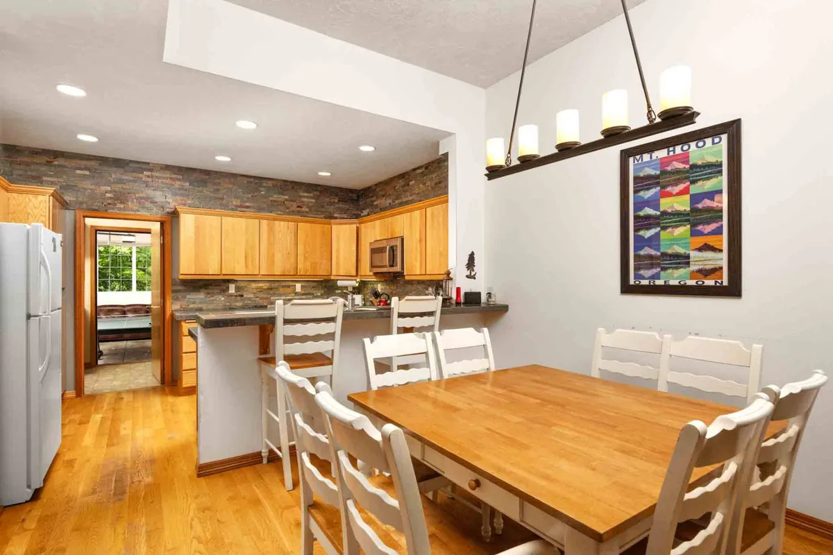 The image showcases a large wooden dining table under a stylish light fixture, adjacent to the kitchen with visible cabinetry and inviting decor. The setup suggests a welcoming environment for family dinners, casual feasts, or celebratory gatherings.