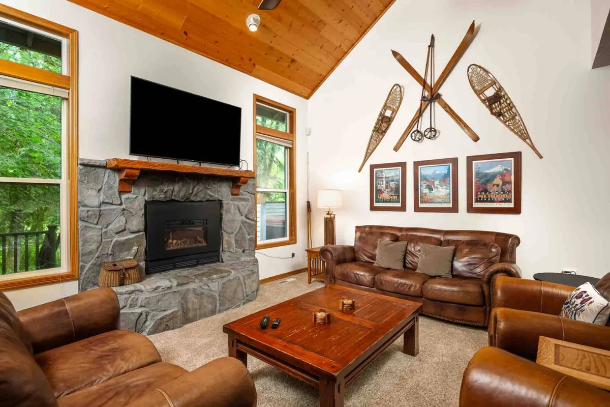 The image showcases a warm, inviting living room centered around a grand stone fireplace, flanked by plush leather sofas and a rustic wooden coffee table. Soft natural light filters in, highlighting decorative wall art and creating a cozy, welcoming ambiance ideal for relaxing after outdoor adventures.