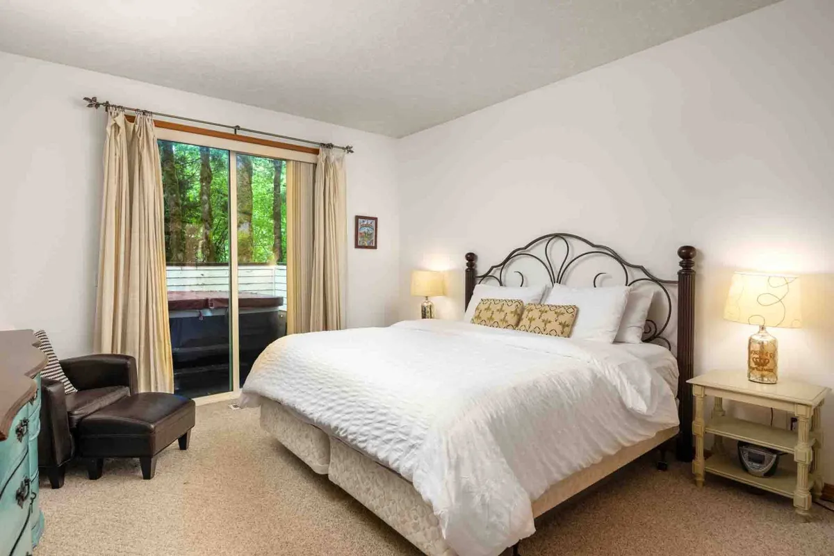 A serene bedroom featuring a neatly made bed with crisp white linens, flanked by nightstands with lamps. A large window with curtains allows natural light to fill the room, creating a bright, calming atmosphere ideal for rest and relaxation.