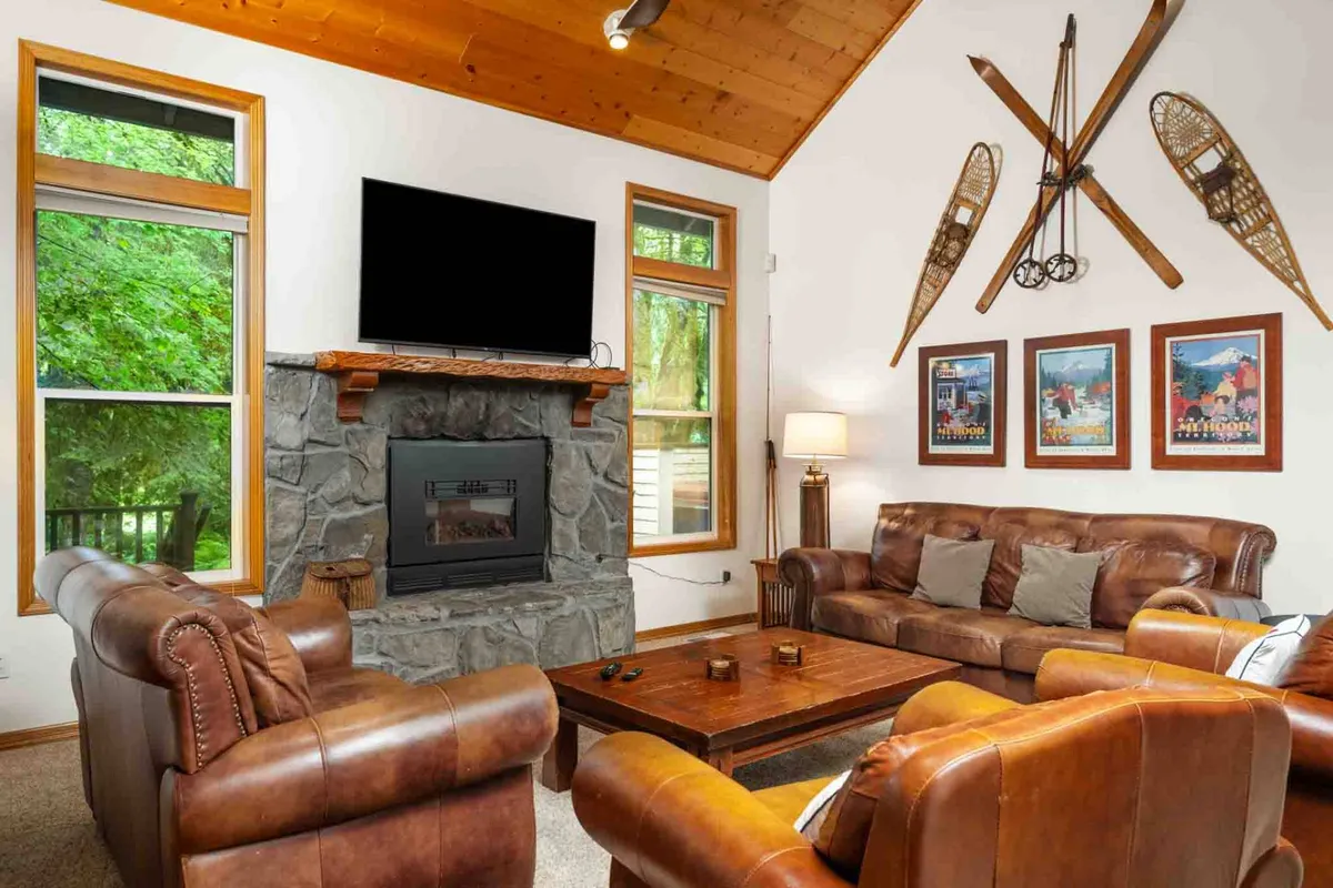 This image showcases a spacious living room with stone fireplace, plush leather sofas, a wooden coffee table, and expansive windows letting in abundant natural light. Decorative Mt. Hood-themed wall art adds a touch of local character and mountain charm to the ambiance. The arrangement of furniture invites relaxation, socializing, and movie nights, embodying a welcoming, open-concept interior.