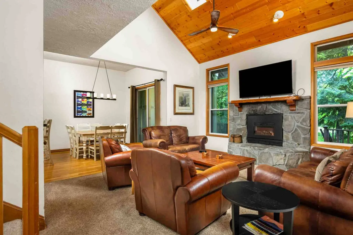 A cozy living area featuring leather armchairs grouped around a stone fireplace and a wooden dining table, with large windows offering serene views. The ceiling fan and warm lighting evoke a sense of comfort, while the open layout connects living and dining spaces, supporting seamless conversation and connection.