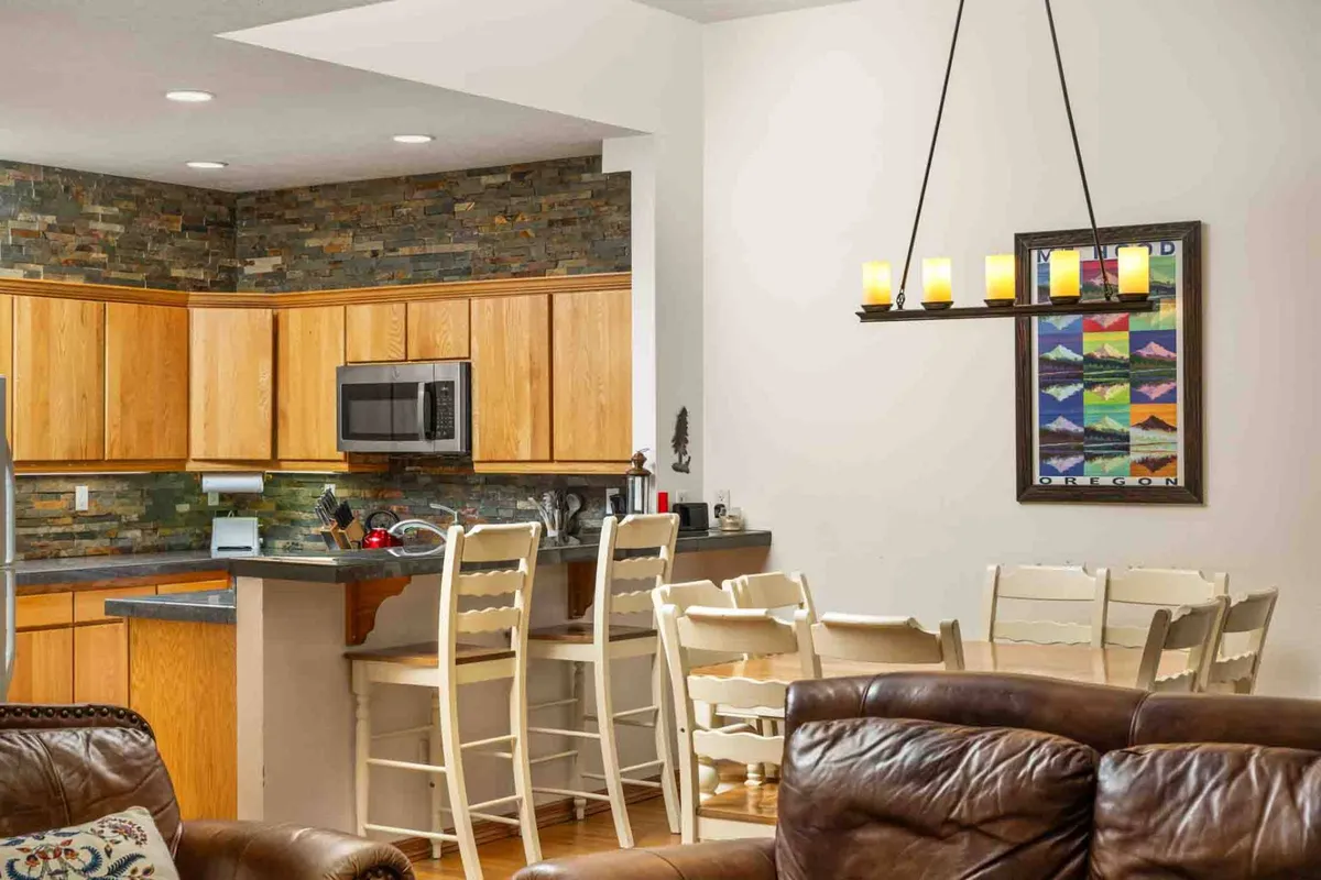 This scene captures the seamless flow between the kitchen and dining areas. It features wooden cabinets, an extended stone backsplash, bar stools at the breakfast bar, and a spacious dining table, set against a cozy backdrop with framed artwork. The arrangement hints at conversations over meals and casual family gatherings.