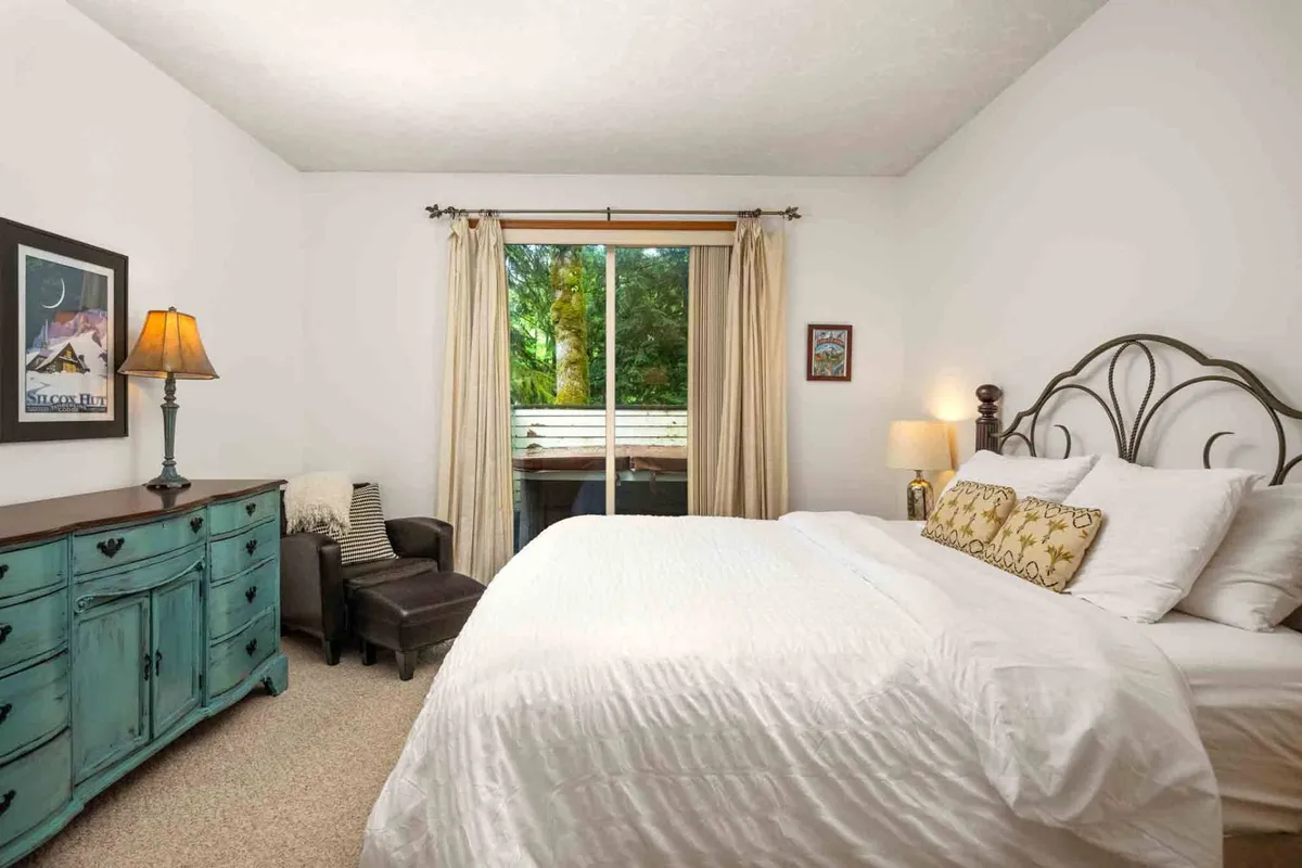 A tranquil queen bedroom with a bed adorned with decorative pillows, a blue vintage dresser, and a window with curtains. The space feels welcoming and cozy, ideal for moments of solitude or quiet rest.